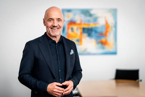 Mann steht in einem schwarzen Hemd vor einer Wand.